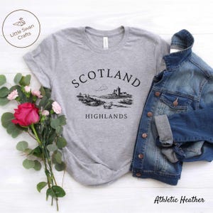 May include: Grey t-shirt featuring a black illustration of a Scottish scene with a castle and sheep, and the words "SCOTLAND HIGHLANDS". A denim jacket is placed beside the shirt. A bouquet of flowers is on the left. The shirt is labelled "Athletic Heather".