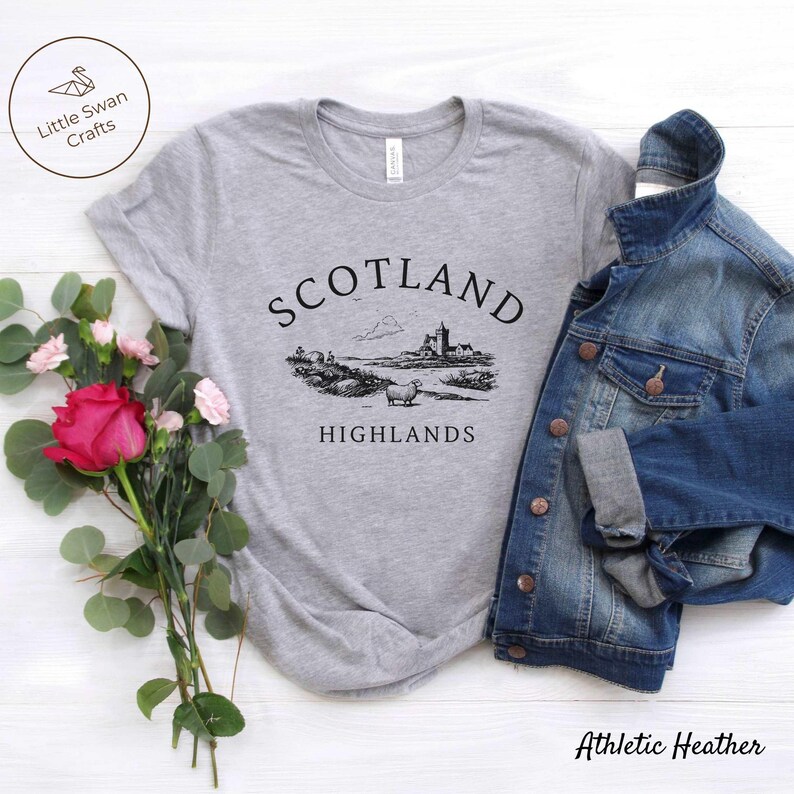 May include: Grey t-shirt featuring a black illustration of a Scottish scene with a castle and sheep, and the words "SCOTLAND HIGHLANDS". A denim jacket is placed beside the shirt. A bouquet of flowers is on the left. The shirt is labelled "Athletic Heather".