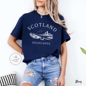 May include: Navy blue t-shirt with a white graphic depicting the Scottish Highlands and the words "SCOTLAND HIGHLANDS". The shirt has a crew neck and short sleeves. The brand logo "Little Swan Crafts" is visible on the left side.