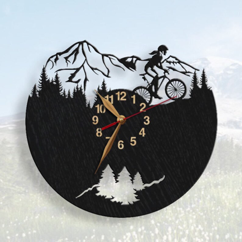 May include: Black circular wall clock featuring a mountain biking scene. The clock has a silhouette design with a cyclist, mountains, and trees. The clock hands are gold with a red second hand, and the numbers are gold.