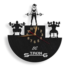 May include: Black and gold clock with a silhouette of two people lifting weights. The clock face has Roman numerals and the words "BE STRONG" are printed below the clock face.