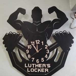 May include: Black wall clock shaped like a weightlifter holding a barbell. The clock face has gold-colored hands and numbers. The text "Luther's Locker" is printed below the clock face.