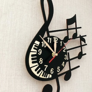 Puede incluir: Reloj de pared negro con forma de clave de sol con notas musicales. La esfera del reloj tiene un dise&ntilde;o de teclas de piano y n&uacute;meros dorados.