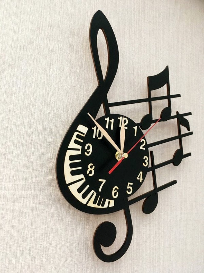 Puede incluir: Reloj de pared negro con forma de clave de sol con notas musicales. La esfera del reloj tiene un dise&ntilde;o de teclas de piano y n&uacute;meros dorados.