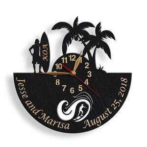 Pu&ograve; includere: Orologio da parete in legno nero a tema surf. Presenta una silhouette di un surfista, palme e una tavola da surf con "KOA". Il quadrante dell'orologio ha lancette e numeri dorati, con i nomi "Jesse and Marisa" e la data "25 agosto 2018".