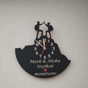 May include: Black clock with a mountain silhouette and two figures high-fiving on top. The clock face has gold numbers and hands. The clock is personalized with the names "Mark & Alisha Stephan" and the word "mountains" with a red heart.