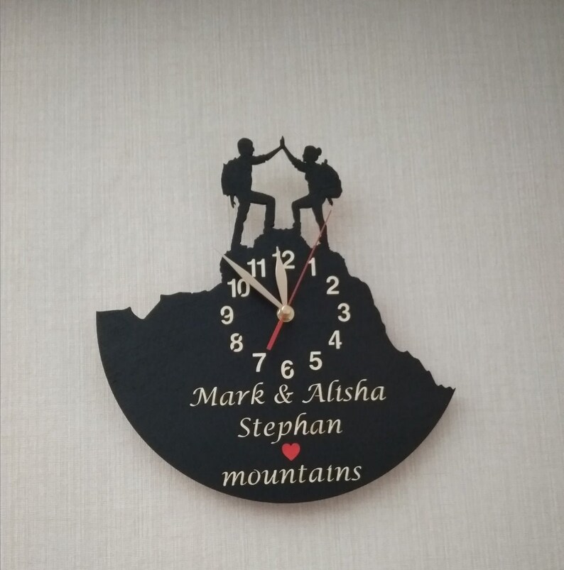 May include: Black clock with a mountain silhouette and two figures high-fiving on top. The clock face has gold numbers and hands. The clock is personalized with the names "Mark & Alisha Stephan" and the word "mountains" with a red heart.