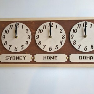 May include: Wooden wall clock with three circular faces displaying different time zones. The clock features the text "SYDNEY", "HOME", and "DOHA" below each clock face. The clock has a light wood frame and a dark wood backing.