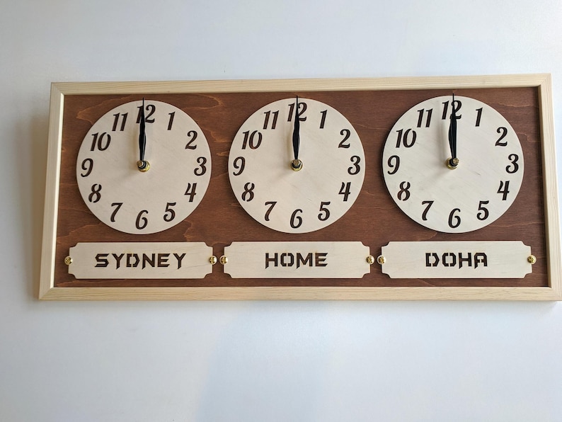May include: Wooden wall clock with three circular faces displaying different time zones. The clock features the text "SYDNEY", "HOME", and "DOHA" below each clock face. The clock has a light wood frame and a dark wood backing.
