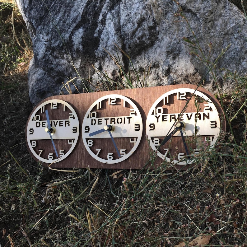 May include: A horizontal wooden wall clock featuring three circular clock faces mounted on a rectangular base. Each face displays the city names Denver, Detroit, and Yerevan in black lettering, with black clock hands and numerical markers.