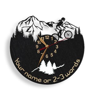 May include: Black circular wall clock featuring a mountain biking scene. The clock has a black background with a mountain range, trees, and a cyclist. The clock hands are gold, and the numbers are gold. Text at the bottom reads "Your name or 2-3 words."