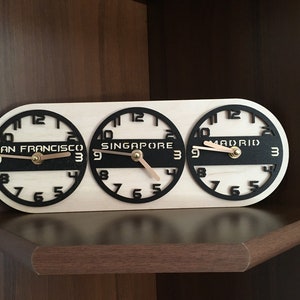 May include: Wooden world time clock displaying the time in San Francisco, Singapore, and Madrid. The clock has a light wood base with black clock faces and hands. The cities' names are printed in a bold font.