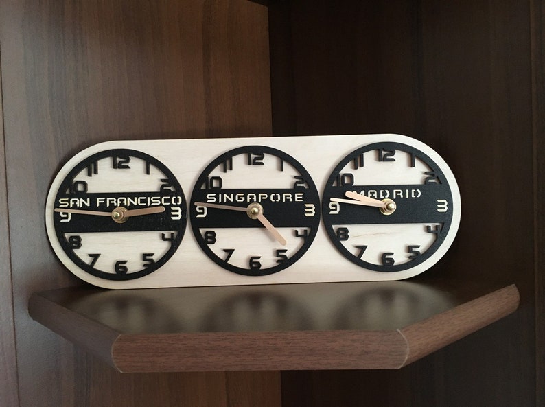 May include: Wooden world time clock displaying the time in San Francisco, Singapore, and Madrid. The clock has a light wood base with black clock faces and hands. The cities' names are printed in a bold font.