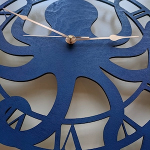 May include: A navy blue wooden wall clock with an octopus design. The clock has copper-coloured hands and Roman numeral hour markers. The octopus is the central design element, with its tentacles forming the clock's face.