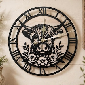 May include: A round wall clock featuring a black silhouette of a Highland cow's face, set against a light wood background. The clock has black Roman numerals and hands. Floral and leaf details are below the cow's face.