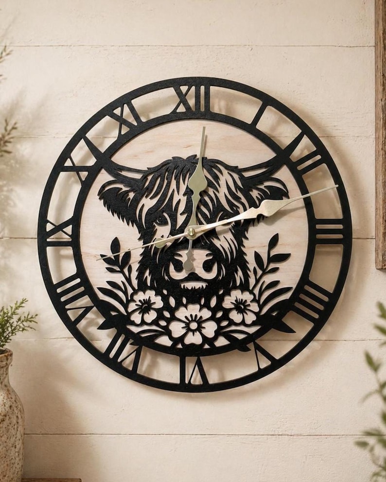 May include: A round wall clock featuring a black silhouette of a Highland cow's face, set against a light wood background. The clock has black Roman numerals and hands. Floral and leaf details are below the cow's face.
