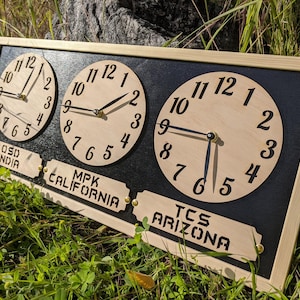 May include: Wooden wall clock with three circular faces displaying different times. The clock has a black background and a light wood frame. Each clock face has black numerals and hands. Labels below read "OSD INDIA", "MPK CALIFORNIA", and "TCS ARIZONA".