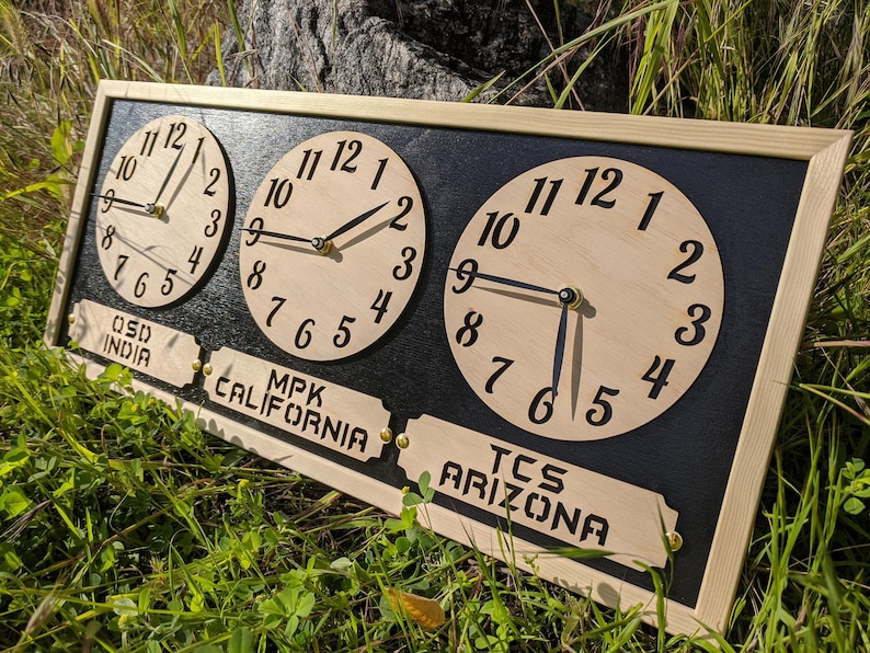 May include: Wooden wall clock with three circular faces displaying different times. The clock has a black background and a light wood frame. Each clock face has black numerals and hands. Labels below read "OSD INDIA", "MPK CALIFORNIA", and "TCS ARIZONA".