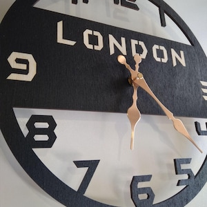 May include: A round, black wooden clock with the word "LONDON" in stencil-style lettering. The clock hands are copper-colored, and the numbers are cut out of the black face. The clock is mounted on a white wall.