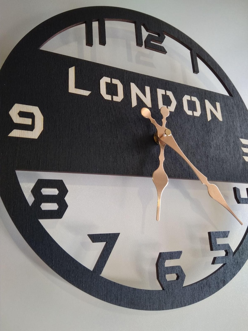 Pu&ograve; includere: Un orologio rotondo in legno nero con la scritta "LONDON" in stile stencil. Le lancette dell'orologio sono color rame e i numeri sono ritagliati. L'orologio &egrave; montato su una parete bianca.