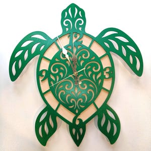 May include: A green wooden turtle-shaped wall clock. The clock features a detailed, openwork design with a light-colored wood backing. The clock hands are silver and gold. The clock is designed to be mounted on a wall.