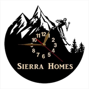 Puede incluir: Reloj en blanco y negro con una escena de monta&ntilde;a y un esquiador. La esfera del reloj tiene n&uacute;meros y manecillas dorados. El texto "SIERRA HOMES" est&aacute; en la parte inferior del reloj.