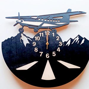 May include: Black wooden wall clock featuring a silhouette of an aircraft flying over mountains. The clock face has white numbers and a gold-coloured hand.