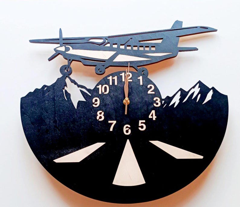 May include: Black wooden wall clock with a silhouette of a plane flying over mountains. The clock face has white numbers and a gold-colored hand.