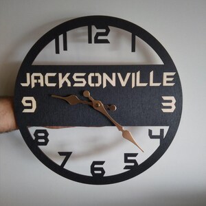 Pu&ograve; includere: Un orologio rotondo in legno nero con il nome "JACKSONVILLE" in un colore chiaro al centro. Le lancette dell'orologio sono color rame e i numeri sono ritagliati dal quadrante nero. L'orologio &egrave; tenuto da una mano.