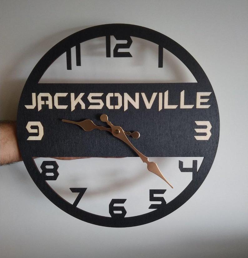 Pu&ograve; includere: Un orologio rotondo in legno nero con il nome "JACKSONVILLE" in un colore chiaro al centro. Le lancette dell'orologio sono color rame e i numeri sono ritagliati dal quadrante nero. L'orologio &egrave; tenuto da una mano.