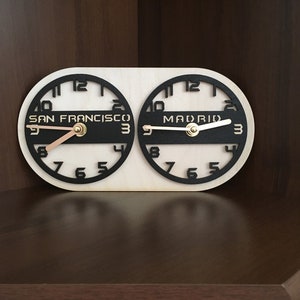 May include: A dual-dial wall clock on a light wood oval base featuring two black circular clock faces. The left dial is labeled San Francisco and the right dial is labeled Madrid. Both clocks have black numbers and metallic gold-colored hands.