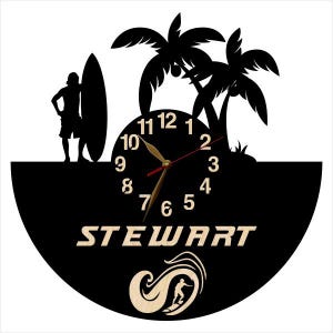 May include: A black wall clock featuring a silhouette of a person standing with a surfboard next to palm trees. The clock face has light-colored numbers, the name STEWART in capital letters, and a graphic of a person surfing inside a wave.