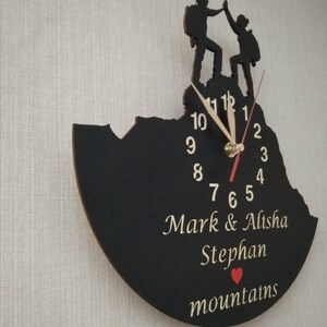May include: Black wooden clock with a silhouette of two people hiking on a mountain. The clock face has white numbers and hands. The clock is personalized with the names "Mark & Alisha Stephan" and the word "mountains" with a red heart.