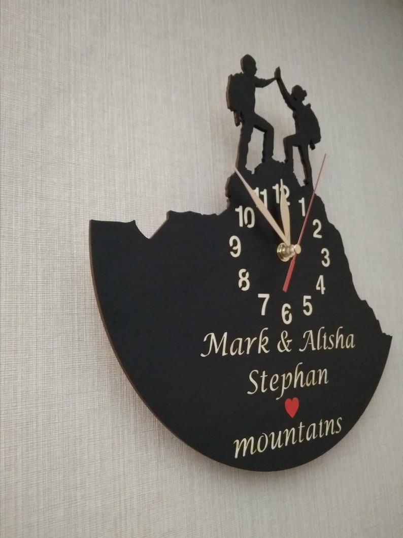 May include: Black wooden clock with a silhouette of two people hiking on a mountain. The clock face has white numbers and hands. The clock is personalized with the names "Mark & Alisha Stephan" and the word "mountains" with a red heart.