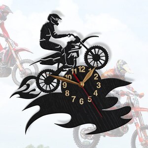 May include: Black wooden wall clock featuring a silhouette of a motocross rider jumping over flames. The clock face has black Roman numeral hour markers and gold hands.