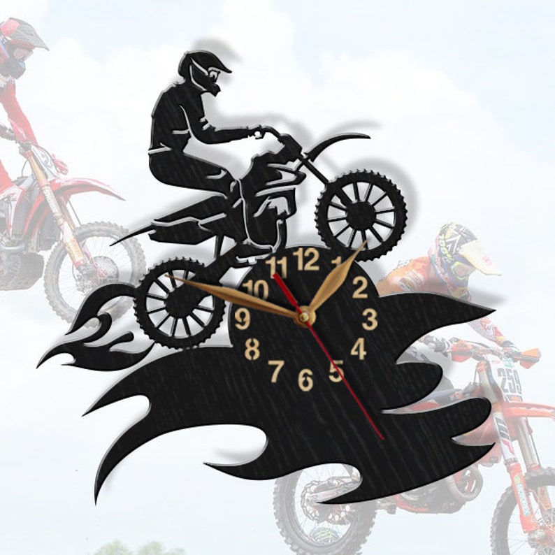 May include: Black wooden wall clock featuring a silhouette of a motocross rider jumping over flames. The clock face has black Roman numeral hour markers and gold hands.