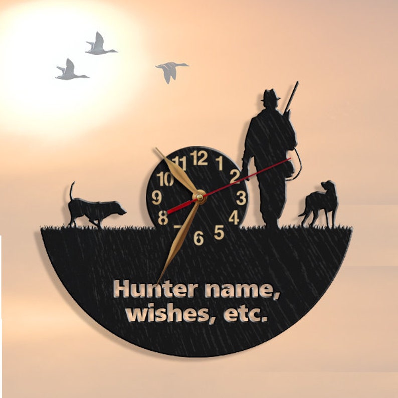 May include: Black wooden clock with a silhouette of a hunter, two dogs, and flying ducks. The clock face has gold hands and Roman numerals. The text "Hunter name, wishes, etc." is printed below the clock face.
