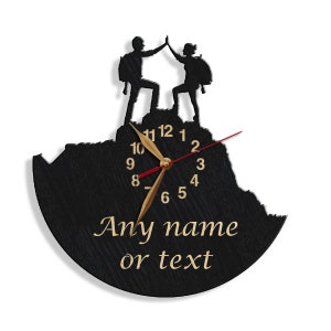 May include: Black wooden clock with a silhouette of two people high-fiving on a mountain top. The clock face has gold hands and black numbers. The text "Any name or text" is printed below the clock face.