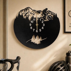 May include: Black metal wall clock featuring a mountain biking scene. The clock has a circular design with a silhouette of a cyclist, trees, and mountains. The clock hands are gold-colored, and the numbers are white.