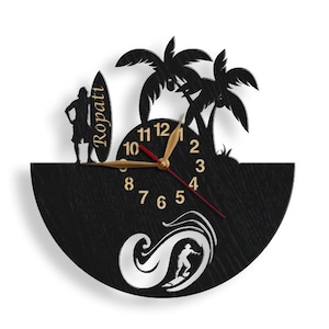 Pu&ograve; includere: Orologio da parete in legno nero a tema surf. L'orologio presenta una silhouette di un surfista, palme e una tavola da surf con la scritta "Ropati". Le lancette dell'orologio sono dorate e rosse.