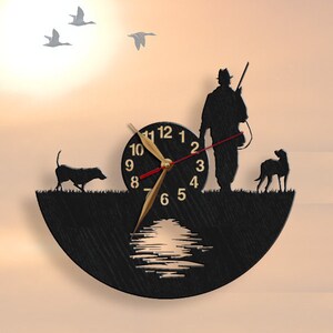 May include: Black wooden wall clock with a silhouette of a hunter, two dogs, and ducks flying overhead. The clock face has gold numbers and hands, and a red second hand. The clock is shaped like a half-circle and has a cutout of a lake with a reflection of the sun.