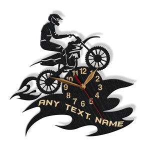 May include: Black and gold wall clock featuring a silhouette of a motocross rider jumping over flames. The clock face has black Roman numeral hour markers and gold hands. The clock face also includes the text "ANY TEXT, NAME".