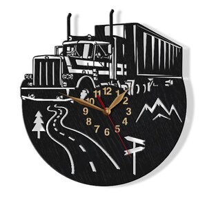 May include: Black and white wall clock featuring a silhouette of a lorry driving along a winding road with mountains in the background. The clock face has gold numbers and hands.