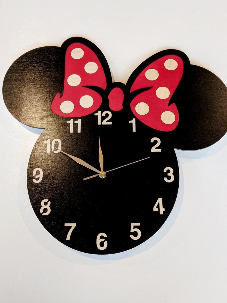 May include: Black wooden clock shaped like a cartoon mouse head with a red bow with white polka dots. The clock face has white numbers 1 through 12 and black hands.