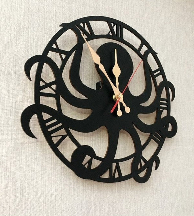 May include: A black octopus wall clock with Roman numerals. The clock has a cut-out design of an octopus in the centre, with the tentacles extending outwards. The clock hands are beige, and the second hand is red. It is mounted on a textured, off-white wall.
