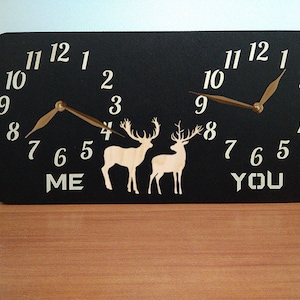 May include: A black wooden clock with two clock faces, one labeled "ME" and the other "YOU". Each clock face has a deer silhouette and gold-colored hands.