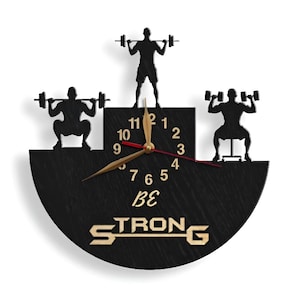 May include: Black and gold wall clock with a half-circle design. The clock features three silhouettes of people lifting weights. The clock face has Roman numerals and the words "Be Strong" are printed below the clock face.