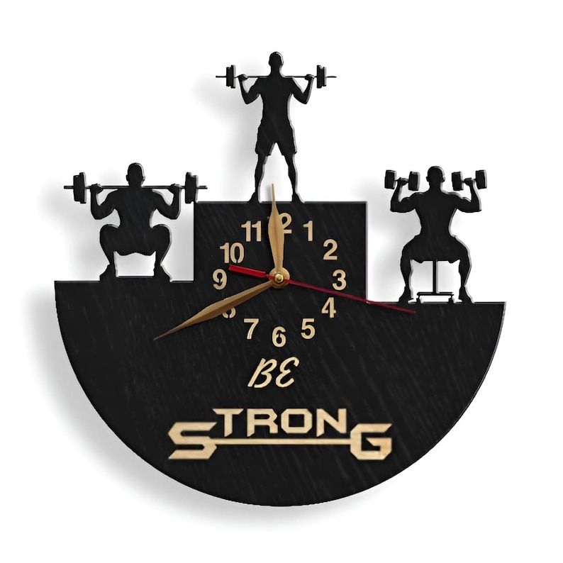 May include: Black and gold wall clock with a half-circle design. The clock features three silhouettes of people lifting weights. The clock face has Roman numerals and the words "Be Strong" are printed below the clock face.
