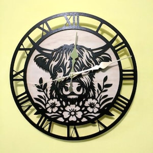 May include: A round wooden wall clock with a black frame and Roman numerals. The clock face features a Highland cow design in black and natural wood tones, with floral accents. The clock hands are gold.