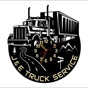 May include: Black and white vinyl clock featuring a silhouette of a semi-truck, mountains, and a winding road. The clock face has gold numbers and hands. The text "J & E TRUCK SERVICE" is printed around the clock face.